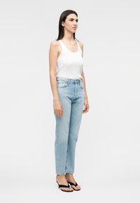 Mujer con cabello largo y oscuro, que lleva una camiseta blanca sin mangas, vaqueros de tiro alto de color azul claro y sandalias negras, está de pie frente a un fondo liso.