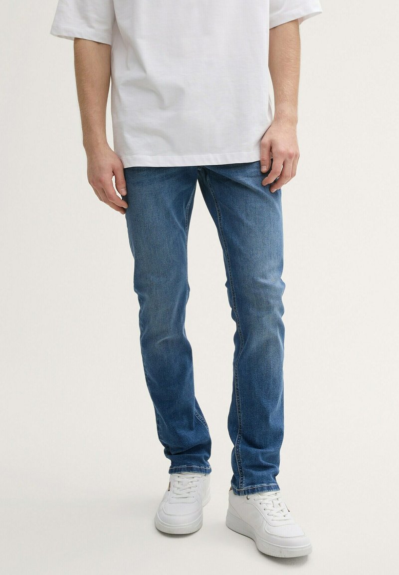 Jeans de mezclilla azul con un ajuste slim, que cuentan con un diseño clásico de cinco bolsillos y acentos de costura, combinados con zapatillas blancas y una camisa blanca.