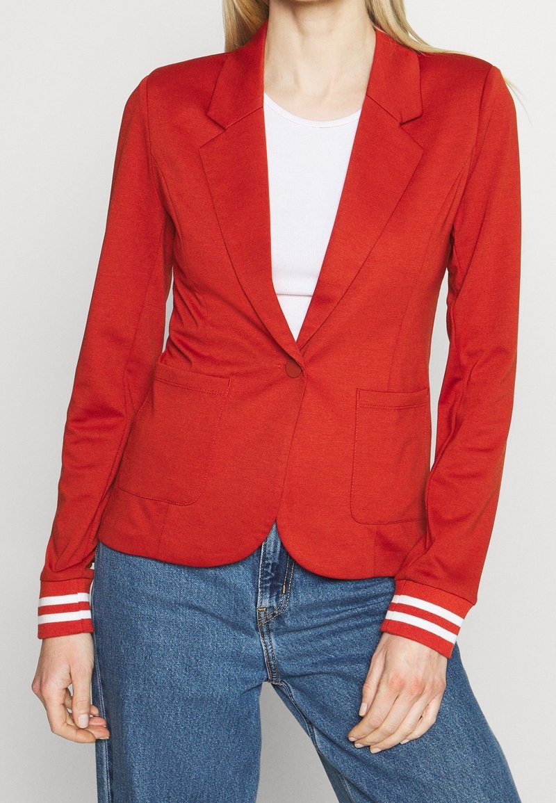 Femme portant un blazer rouge vif avec des poignets rayés, un haut blanc et un jean bleu, debout devant un fond clair et uni.