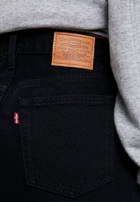 Jean en denim noir avec une étiquette en cuir fauve portant l'inscription « Levi Strauss & Co. » sur la ceinture. Étiquette de marque rouge sur la poche arrière.