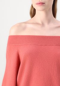 Pull corail décolleté fabriqué à partir d'un tissu doux et texturé, avec un col côtelé et une coupe décontractée, mettant en valeur une surface lisse.