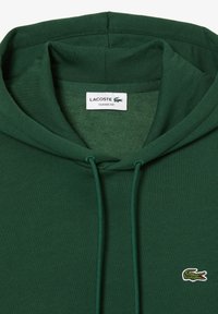 Sudadera verde oscura con cordones, que presenta un pequeño logo bordado de cocodrilo de Lacoste y una etiqueta blanca que dice "Lacoste Classic Fit."