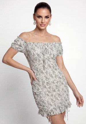 Donna con capelli tirati indietro che indossa orecchini a goccia con perle e un vestito bianco a spalle scoperte con una piccola stampa floreale grigia e orlo arricciato.