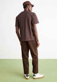 T-shirt en coton marron, coupe décontractée, associé à un pantalon en velours côtelé marron et des baskets blanches. La casquette assortie à la couleur du t-shirt. Design simple, sans motifs.