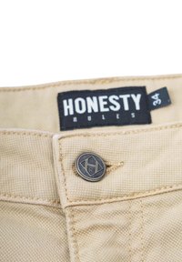 Beige Baumwollhosen verfügen über einen Metallknopf mit geometrischem Design. Ein schwarzes Etikett trägt die Aufschrift "HONESTY RULES" mit der Größe "34".