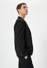 Blazer nero su misura con design a un solo petto, rever a punta, tasche laterali e dettagli con bottoni. Tessuto liscio con una leggera consistenza.