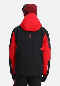 Schwarze und rote Skijacke mit Kapuze, einem Spinnenlogo auf der Rückseite und reflektierenden Akzenten an den Ärmeln. Hergestellt aus wasserabweisendem Material.