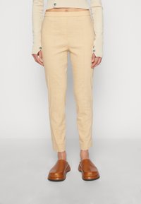 Femme portant un pantalon taille haute beige et des chaussures slip-on marron, se tenant devant un fond blanc uni.