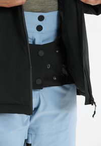 Schwarze Jacke mit glatter Textur, die einen hellblauen Bund mit zwei runden schwarzen Knöpfen und passendem blauen Stoff darunter zeigt.