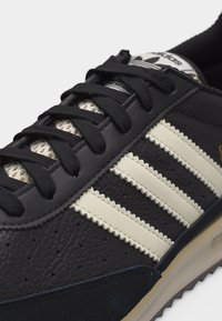 Κοντινό πλάνο μαύρου αθλητικού παπουτσιού Adidas με λευκές, υφασμένες ρίγες, μαύρα κορδόνια και μπεζ σόλα.