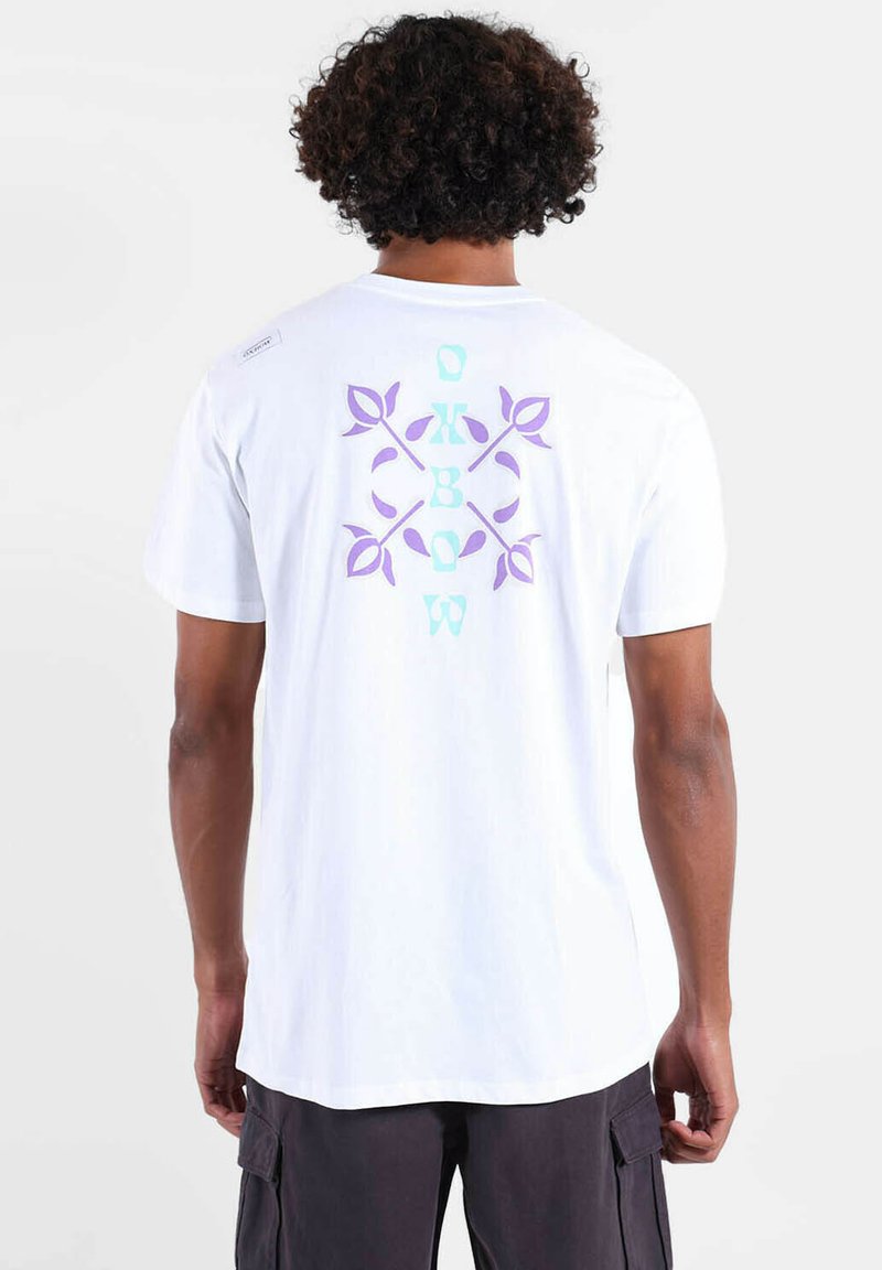 T-shirt blanc avec un motif graphique de feuilles violettes et aqua ainsi qu'un texte au dos. Manches courtes et coupe décontractée. Texture en coton lisse.