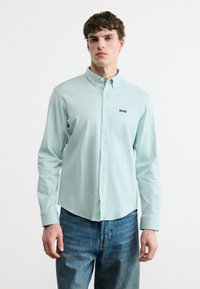 Νεαρός άνδρας με κοντά σγουρά μαλλιά, που φοράει ανοιχτό γαλάζιο πουκάμισο με κουμπιά και λογότυπο "BOSS" και μπλε τζιν, στέκεται μπροστά σε απλό λευκό φόντο.