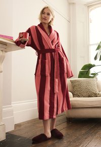 Striped terrycloth bathrobe in pink and burgundy, featuring two front pockets, a tie belt, and soft texture. Model holding a red mug.