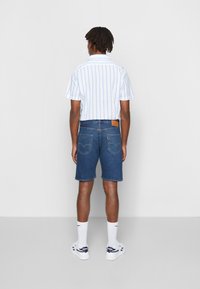 Camisa blanca de manga corta con rayas azules, combinada con pantalones cortos de mezclilla azul oscuro. Calcetines blancos y zapatillas deportivas completan el atuendo.