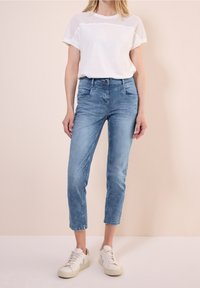 Vit t-shirt med meshdetaljer, kombinerad med ljusblå denim capribyxor och vita sneakers med gröna accenter. Minimalistisk design.