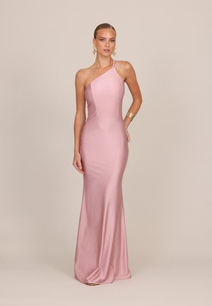 Femme portant une robe rose ajustée longue jusqu'au sol, à une épaule, avec des boucles d'oreilles dorées et un bracelet doré, debout sur un fond uni.