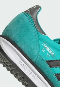 Teal nubuk i tkanina tenisice s crnim trakama i bijelim potplatom, sivi uspon na peti i "adidas SL '72" brend.