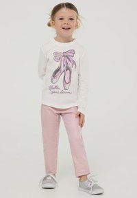 Biała koszula z długim rękawem i falbaniastym kołnierzykiem, z graficznym motywem różowych baletków, w zestawie z jasnoróżowymi spodniami oraz srebrnymi sneakerami z białymi sznurówkami.