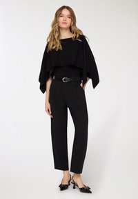 Top nero a strati con maniche ampie, abbinato a pantaloni neri sartoriali e décolleté slingback nere con dettaglio fiocco decorativo.