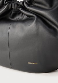 Zwarte zachte leren handtas met samengeplooide bovenkant en klein gouden "Coccinelle"-logo aan de voorkant tegen een witte achtergrond.
