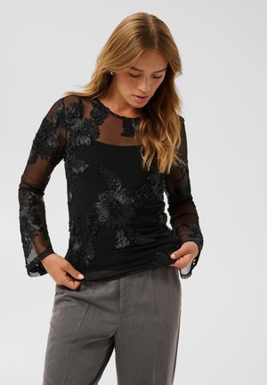 Mulher com cabelo longo e loiro, vestindo um top preto transparente com bordados florais e calças cinzentas, olhando para baixo enquanto ajusta a camisa.