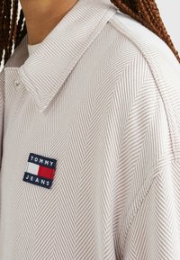 Light pink shirt with a herringbone pattern, featuring a pointed collar and a navy patch labeled "TOMMY JEANS" with red and white accents.