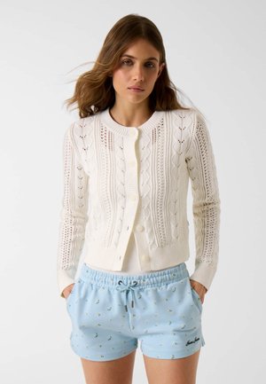 Giovane donna che indossa un cardigan bianco a maglia e pantaloncini con coulisse a fiori azzurri chiaro, in piedi con le mani nelle tasche su sfondo bianco.