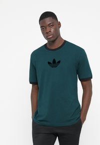 Mørk teal bomulds T-shirt med korte ærmer og sorte ribbede detaljer, der har et sort logo på brystet og en afslappet pasform.