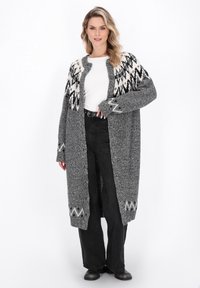 Lange grijze cardigan met zwart-wit gestreepte yoke, open voorkant en een textuur gebreide stof. Gepaard met zwarte broek met wijde pijpen en zwarte laarzen.