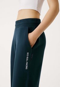 Zbliżenie osoby ubranej w ciemnoniebieskie spodnie dresowe Red Bull Racing z ręką w kieszeni i białym, krótkim topem na jednolitym tle.