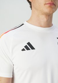 Camiseta deportiva blanca con acentos negros y logo de Adidas. Presenta rayas de colores en rojo, amarillo y negro en el hombro. Mangas cortas.