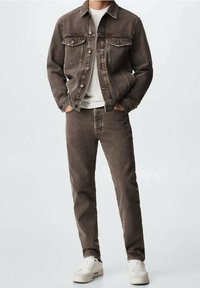 Giacca di jeans marrone con tasche frontali e bottoni, abbinata a jeans marroni coordinati e sneakers bianche. Design semplice e silhouette aderente.