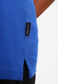 Nära bild av blått tyg som visar en svart Calvin Klein-etikett sydd nära fållen, med en sidospalt på plagg.