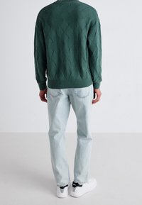 Pull en maille vert émeraude avec motif losange, poignets et ourlet côtelés. Assorti à un jean bleu clair et des baskets blanches avec des accents noirs.