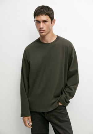 Giovane uomo dai capelli corti e scuri che indossa una camicia verde scuro a maniche lunghe e pantaloni neri, in piedi su uno sfondo bianco.