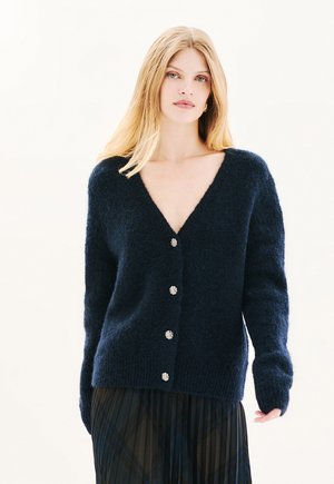 Navyblauwe gebreide cardigan met een V-hals, gestructureerde stof en vijf zilveren bloemenvormige knopen aan de voorkant. Lange mouwen, relaxte pasvorm.