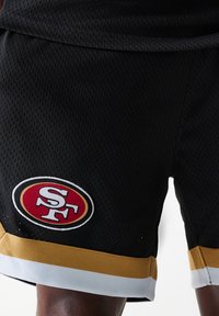 Sorte mesh sportshorts med gull- og hvit trim, med en fremtredende rød og hvit brodert logo på venstre ben.