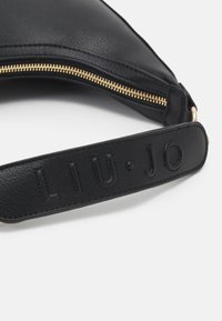 Bandoulière de sac en cuir noir avec le logo "LIU·JO" embossé et une fermeture éclair dorée visible sur le bord du sac sur fond blanc.