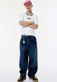 Ung person i hvid skjorte med korte ærmer, løse mørke jeans med blomster-nøglekæde, sorte sko og lyserød kasket, der står med armene korslagt.