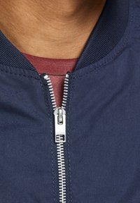 Marinblå bomberjacka med en silverfärgad dragkedja, ribbat krage och en vinröd skjorta under. Tyget är slätt med en subtil glans.