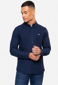 Camisa azul marino de botones con cuello; confeccionada en una tela suave con un ligero brillo. Presenta un pequeño logo en el pecho.