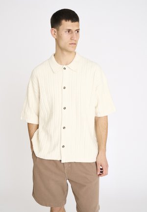 Ung mand med kort mørkt hår iført en cremefarvet kortærmet knaptrøje og lysebrune knælange shorts, hænderne i lommerne.