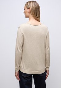 Béžový svetr s dlouhým rukávem a výstřihem do tvaru lodičky vzadu, s žebrovanými manžetami a lemem. Měkká textura, volný střih, kombinováno s tmavě modrými džínami.