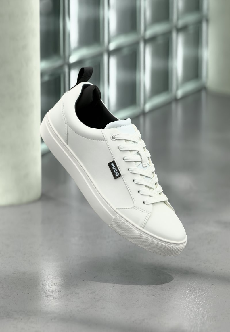 Baskets en cuir blanc avec doublure intérieure noire, fermeture à lacets plats et détail logo visible. Semelle en caoutchouc texturée pour une meilleure adhérence.