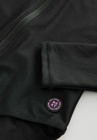 Giacca sportiva nera con colletto arrotondato, chiusura con zip e polsini cuciti. Include una toppa circolare con logo e dettagli rosa.