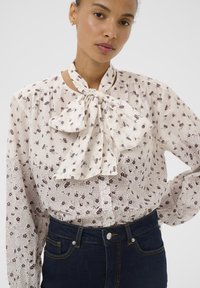 Blusa de manga larga en tela color crema con estampado floral burdeos, con lazo en el cuello, cierre con botones y detalles fruncidos en los hombros.