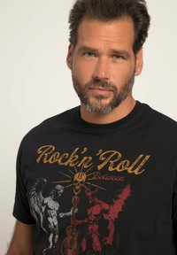 Schwarzes T-Shirt mit einem grafischen Design, das geflügelte Engel- und Teufelsfiguren, einen Schädel und eine Gitarre in metallischen Gold- und Rottönen zeigt.