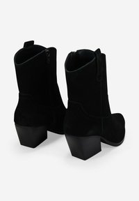 Par de botas de ante negro hasta la mitad de la pantorrilla con tacones cuadrados, cremalleras laterales y lengüetas para tirar, vistas desde la parte trasera sobre un fondo blanco.