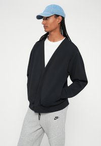 Crna, oversized, dukserica s V-izrezom i dugim rukavima. Uparena s svijetlosivim trenirkama s Nike logom. Model nosi svijetloplavu kapu.