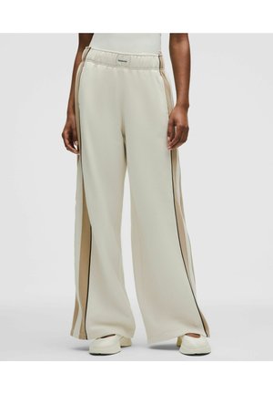 Kremfarvede bukser med vide ben og beige striber på siden, båret med off-white slip-on sko på lys baggrund.